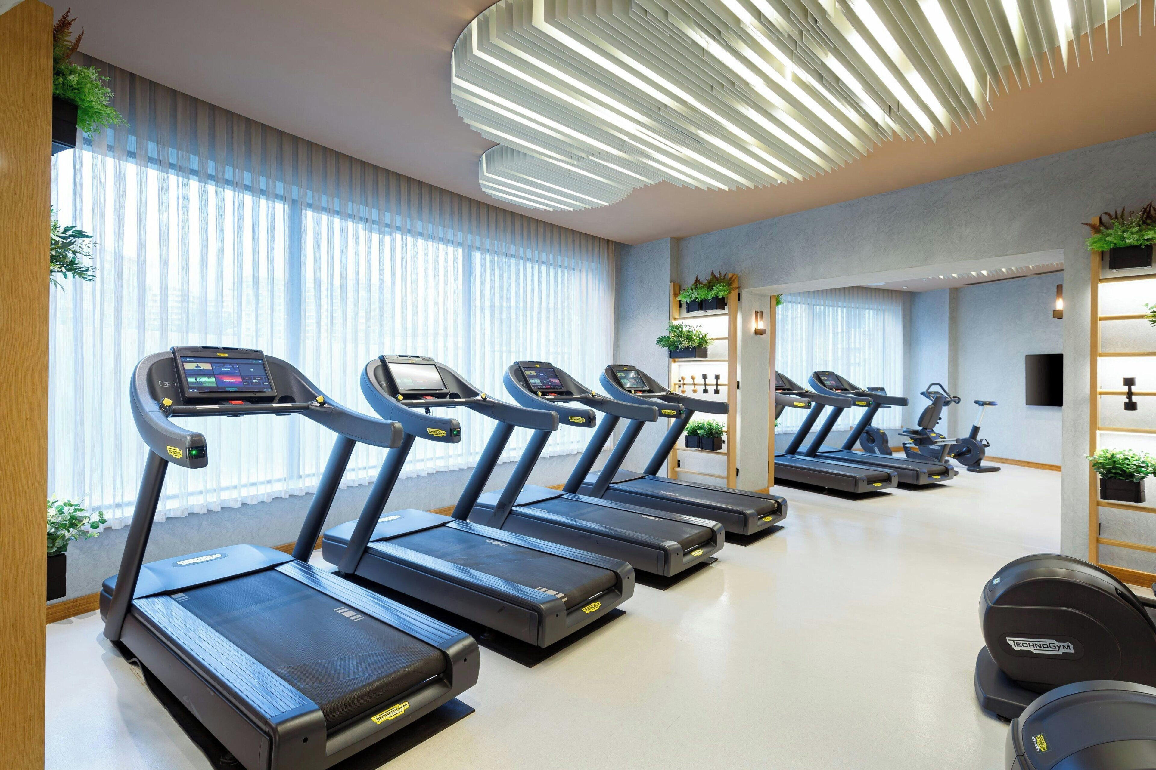 Ein Fitnessraum mit mehreren Laufbändern an einer Wand mit großen Fenstern, einer Decke mit einer modernen Leuchte und einem Spiegel, der den Fitnessbereich reflektiert.