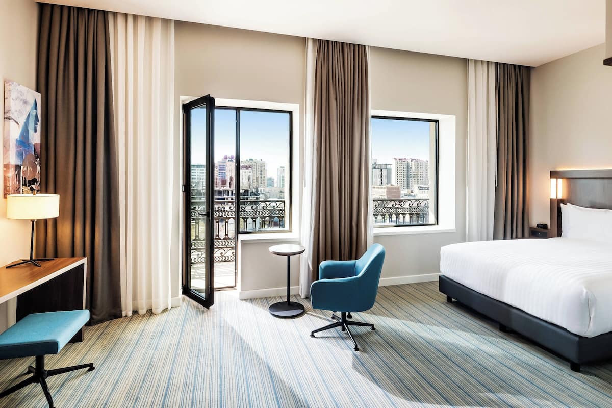 A hotel room with a large bed, a desk with a chair, a small round table, and a view of the city through large windows.