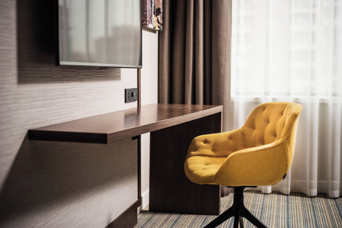 A hotel room with a wooden desk, a yellow armchair, a flat-screen TV mounted on the wall, and a window with curtains.
