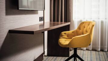 A hotel room with a wooden desk, a yellow armchair, a flat-screen TV mounted on the wall, and a window with curtains.