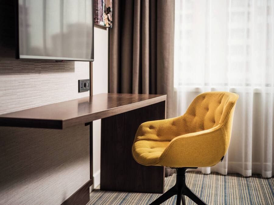 A hotel room with a wooden desk, a yellow armchair, a flat-screen TV mounted on the wall, and a window with curtains.
