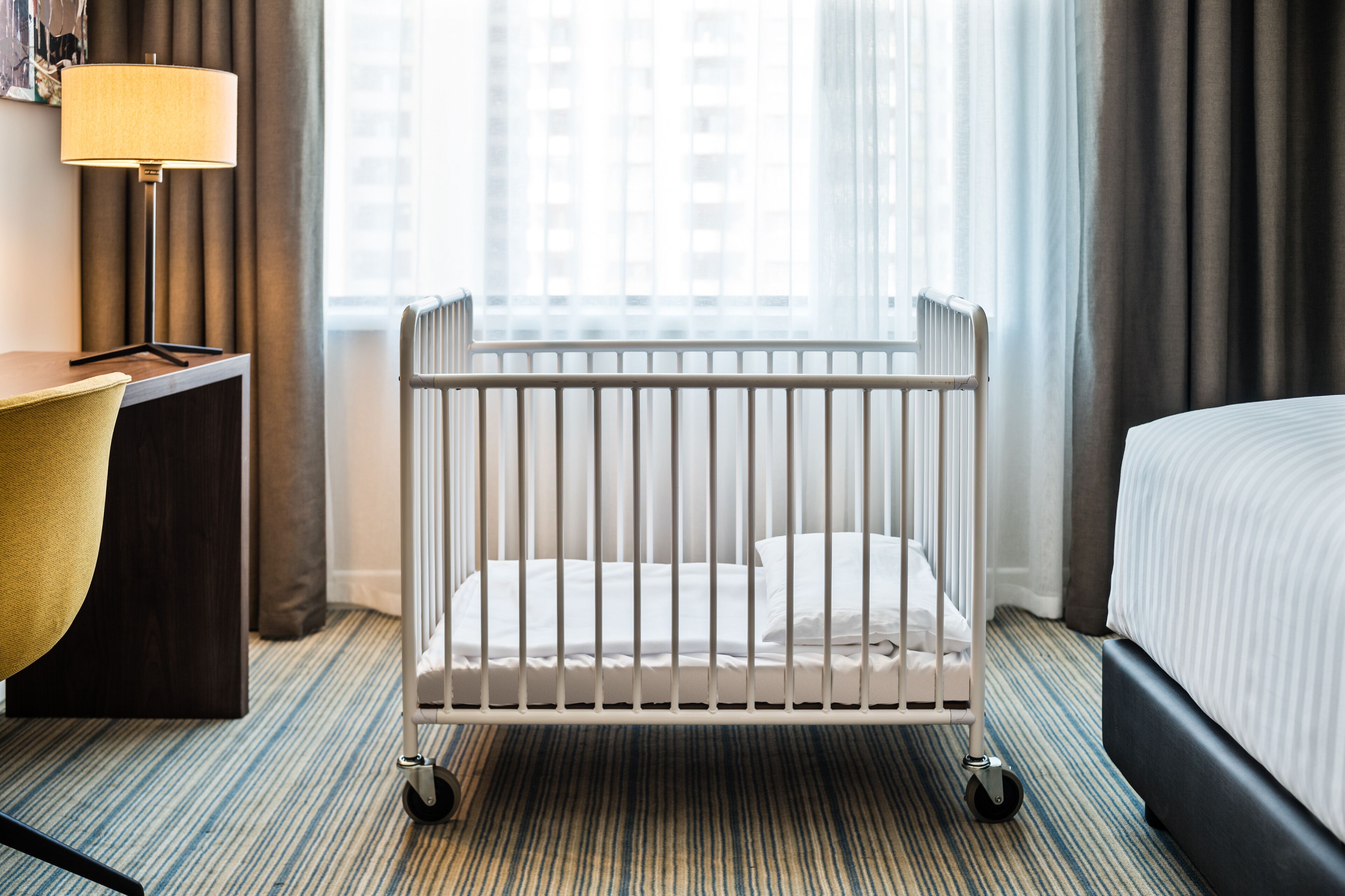 A hotel room with a crib, a desk with a lamp, a bed, and a view of buildings through sheer curtains.