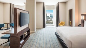 Habitación de hotel con una cama grande, televisor, escritorio y vista a la ciudad.