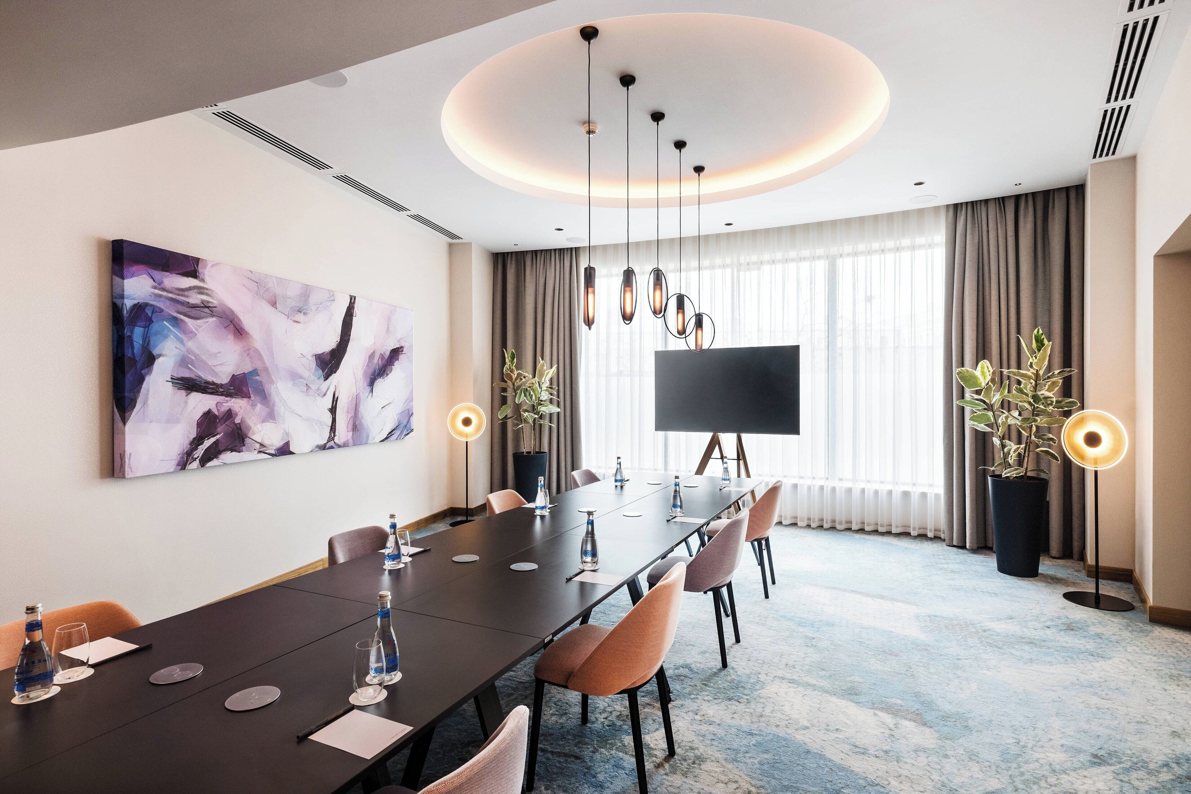 A modern conference room with a long table, chairs, a large painting, a TV, and potted plants.