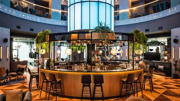 Un moderno vestíbulo de hotel con un bar central, varias zonas de estar y un piso con diseño geométrico.