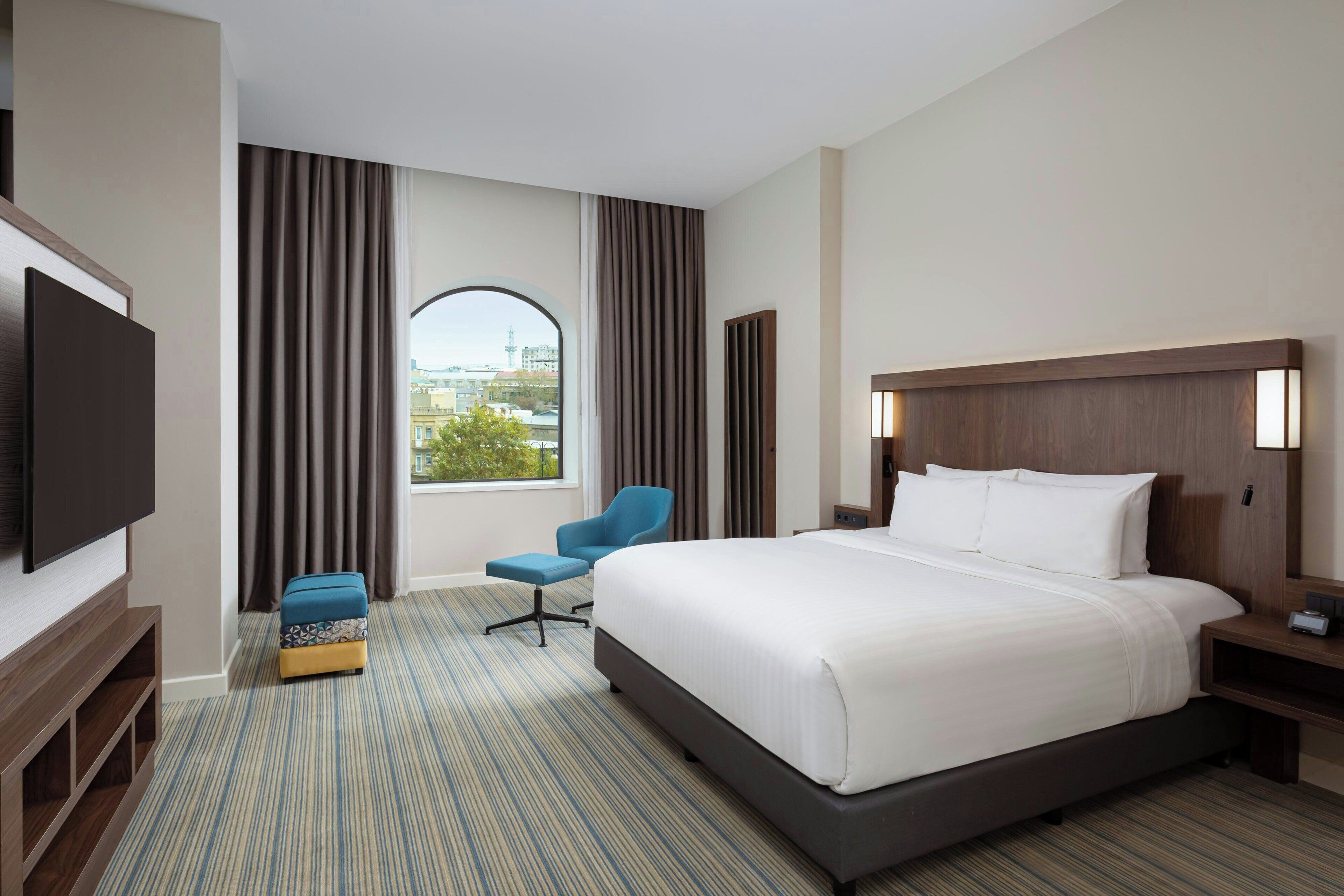 A hotel room with a large bed, a TV, a small seating area, and a window with a view of the city.