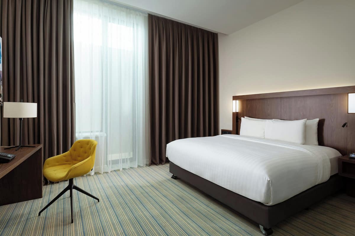 A hotel room with a large bed, a desk with a chair, and a view of the outdoors through sheer curtains.