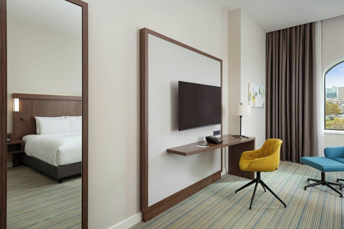 A hotel room with a bed, a TV mounted on a wall, a desk with a chair, and a view of the city through a window.