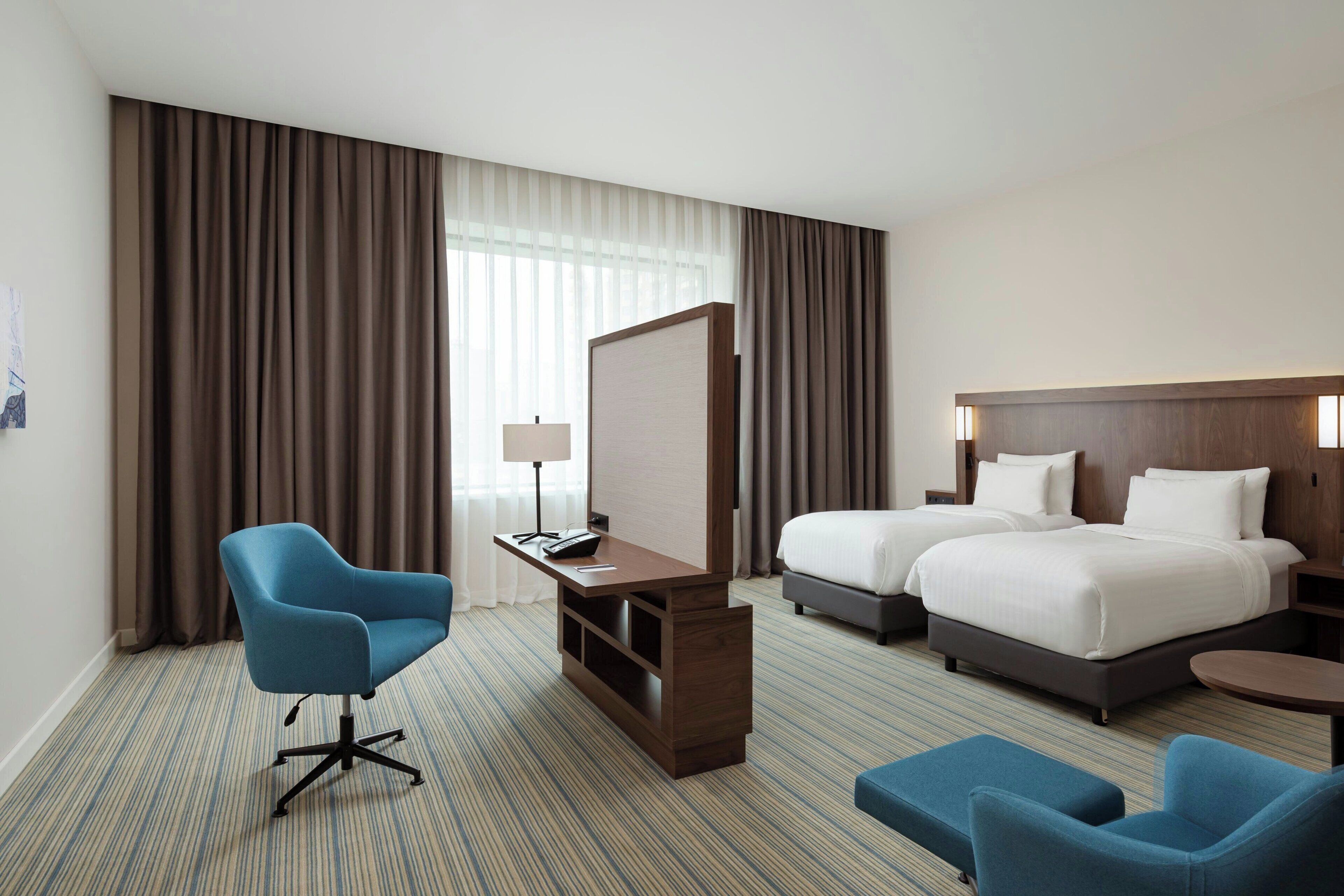 A hotel room with a bed, two blue armchairs, a desk with a lamp, and a large window with curtains.
