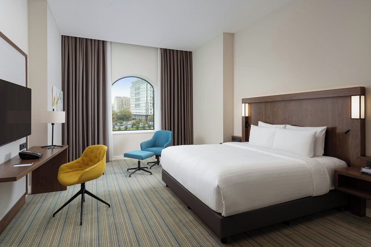 A hotel room with a large bed, a desk with a flat-screen TV, a blue armchair, and a window with curtains.