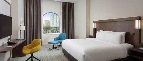 Habitación de hotel con una cama grande, un escritorio con televisor de pantalla plana, un sillón azul y una ventana con cortinas.
