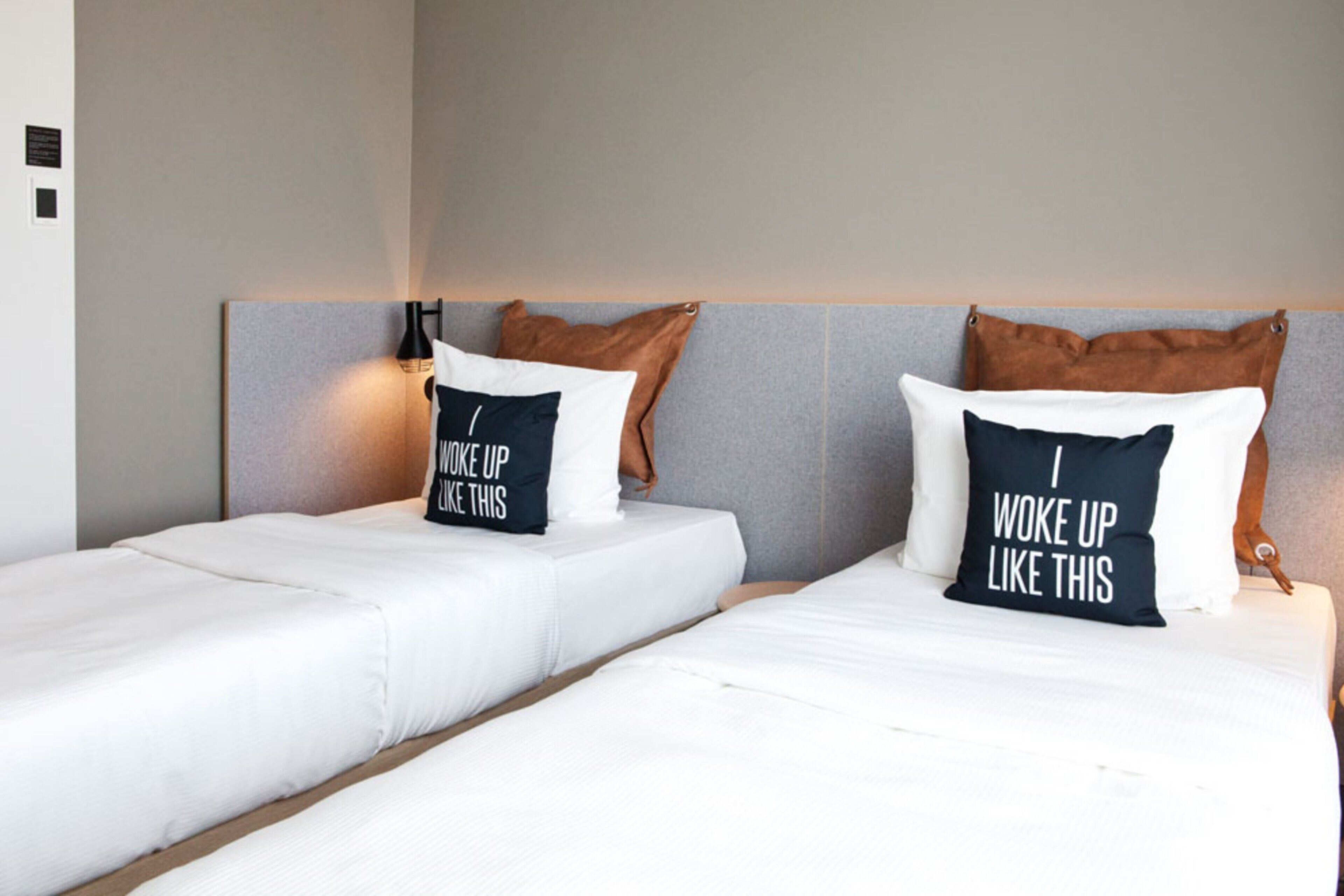 Two single beds with white bedding and pillows with motivational text, a headboard, and a wall-mounted lamp.