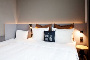 A neatly made bed with white linens and a pillow with the text "I woke up like this".