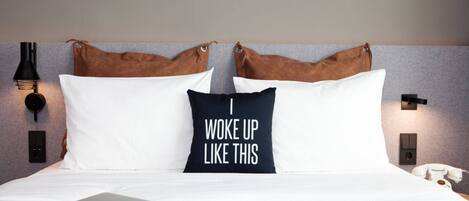 Una cama bien hecha con un cojín que dice “Me levanté así”, una computadora portátil y un teléfono sobre la cama.