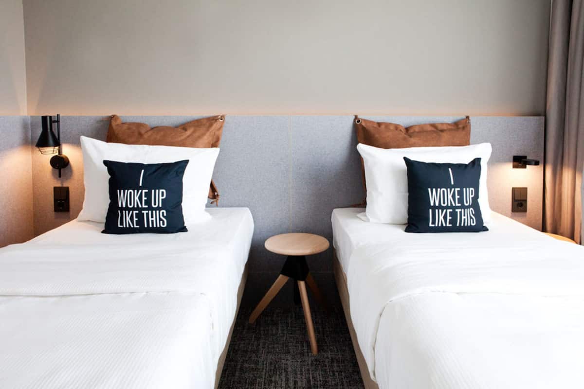 Une chambre d’hôtel avec deux lits, chacun agrémenté d’un oreiller sur lequel est brodé le message « JE ME SUIS RÉVEILLÉ(E) COMME ÇA ». Une petite table ronde se trouve entre les deux lits, et la chambre est décorée dans des tons neutres, avec un mur gris et un plancher en bois.