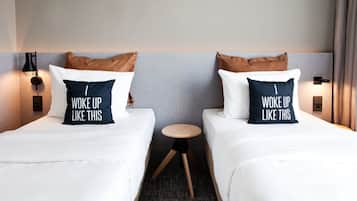 A hotel room with two beds, each with a pillow that reads "WOKE UP LIKE THIS." There is a small round table between the beds, and the room has a neutral color scheme with a grey wall and a wooden floor.