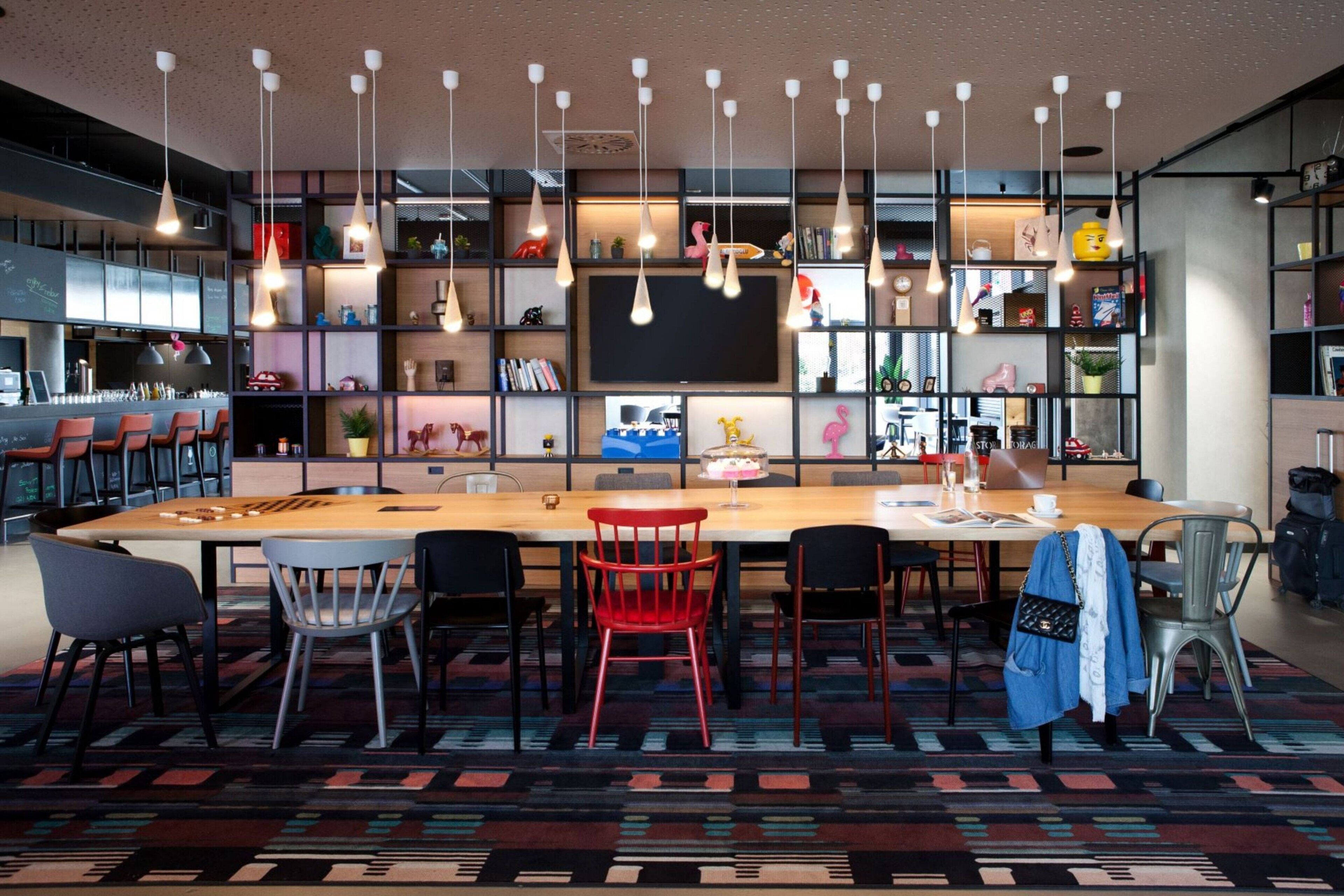 A modern bar area with a long wooden table, multiple chairs, hanging pendant lights, and a variety of decorative items on shelves.