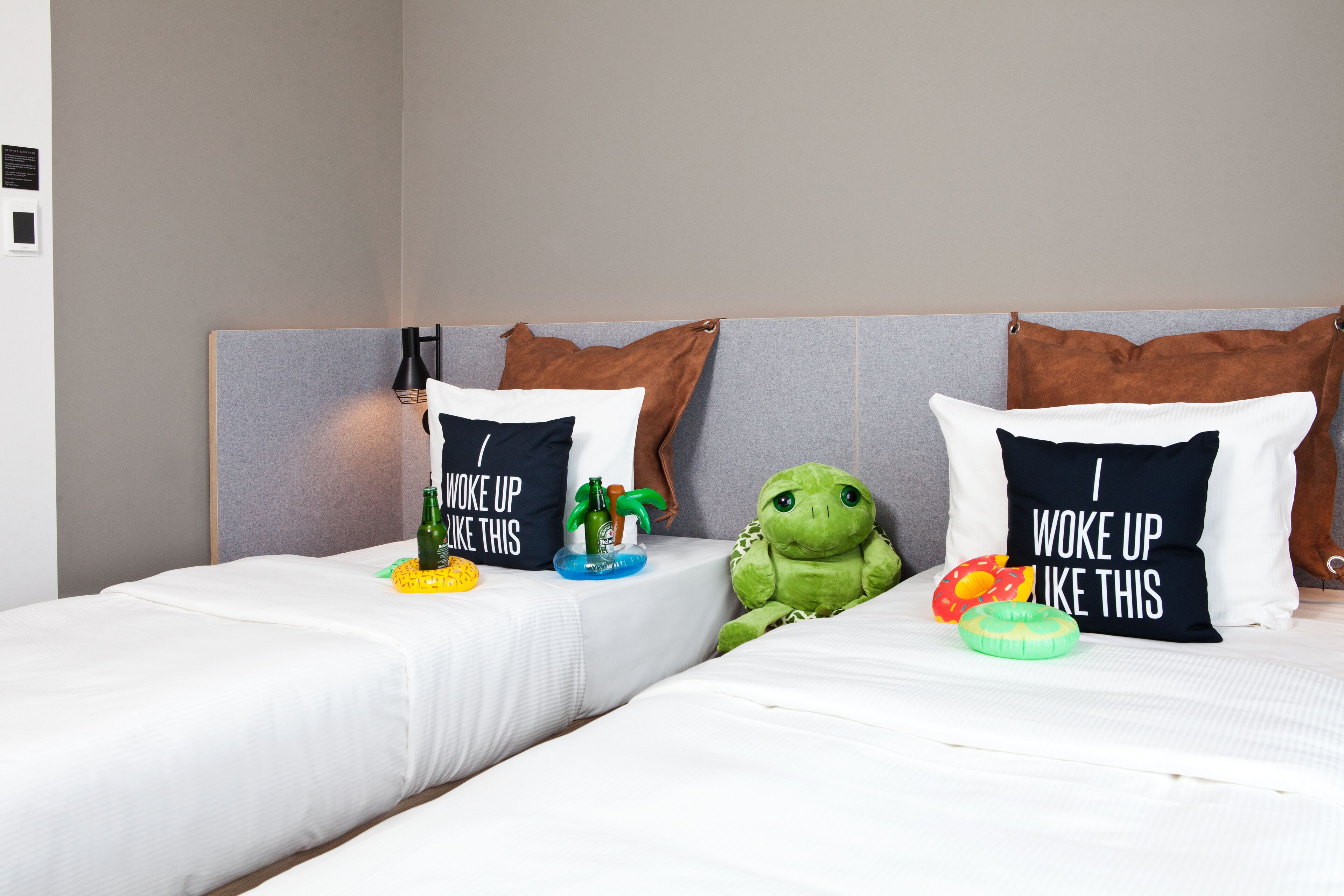 Duas camas de solteiro com roupa de cama branca, cada uma com uma almofada com texto divertido. Um brinquedo de pelúcia verde está colocado entre as camas.