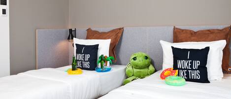 Duas camas de solteiro com roupa de cama branca, cada uma com uma almofada com texto divertido. Um brinquedo de pelúcia verde está colocado entre as camas.