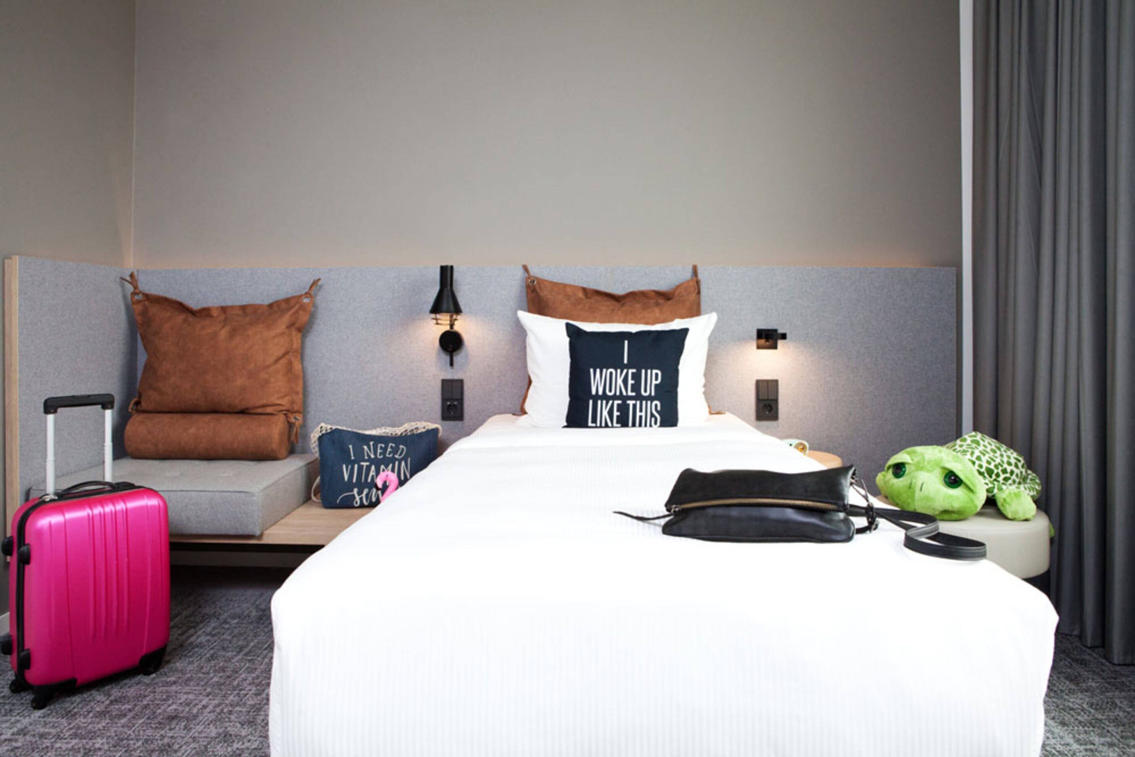 Una habitación de hotel con cama, almohadas, una maleta, una tortuga de peluche y una lámpara fijada a la pared.