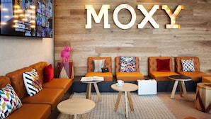 Un moderno espacio lounge con un gran letrero iluminado que dice 'MOXY', asientos cómodos y una pantalla digital mostrando imágenes.