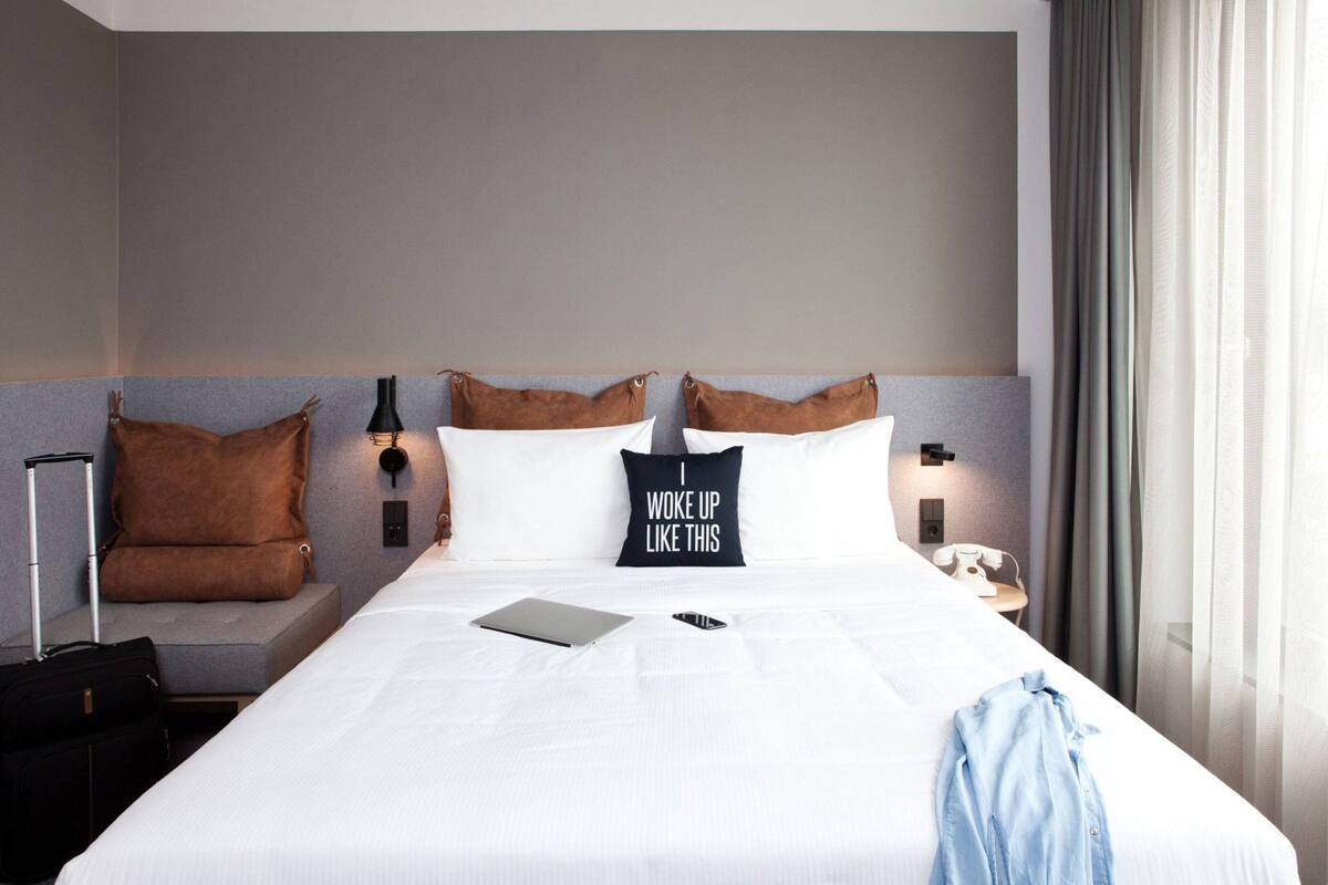 Une chambre d’hôtel avec un grand lit, une table de chevet, une lampe, une valise et un oreiller orné d’un motif.