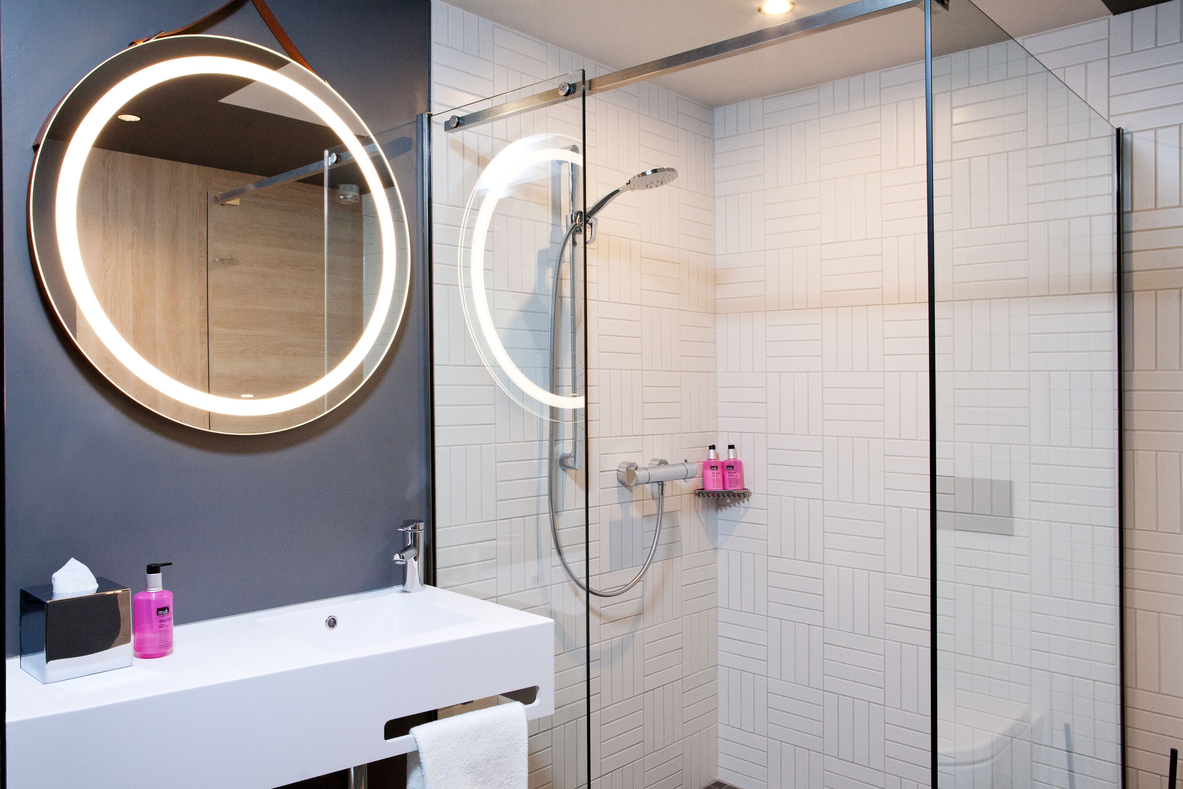 Un baño moderno con ducha envuelta en vidrio, un espejo redondo con luz y un lavamanos blanco con artículos de aseo.