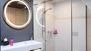 Un baño moderno con ducha envuelta en vidrio, un espejo redondo con luz y un lavamanos blanco con artículos de aseo.