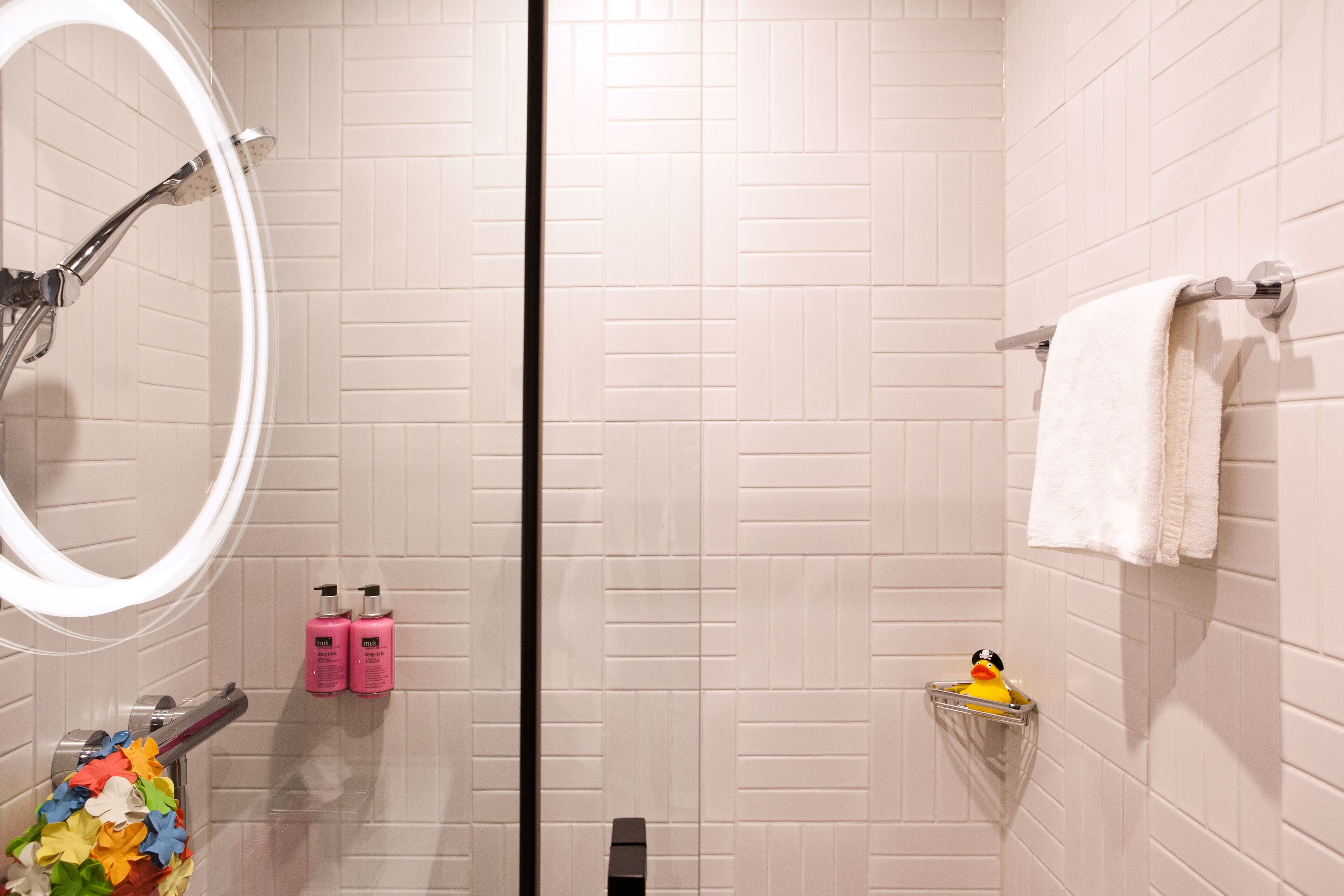 A bathroom with white tiled walls, a shower with a glass door, a mirror, a towel rack, and a shelf with a rubber duck.