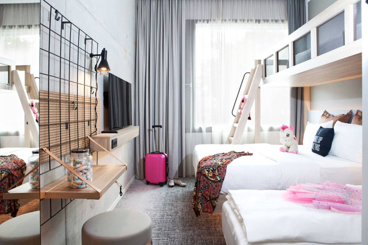 Une chambre d’hôtel avec un lit superposé, un bureau avec une lampe, une valise et un jouet en peluche sur le lit.