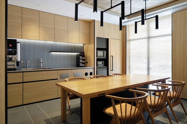A modern kitchen with wooden cabinets, a herringbone backsplash, and a built-in oven. There is a dining table with chairs and pendant lighting.