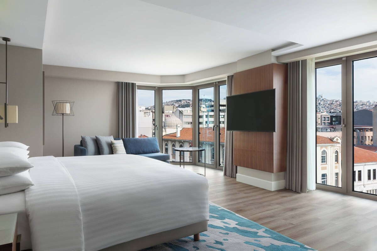 A modern hotel room with a large bed, a flat-screen TV, a small table, and a view of the city through large windows.