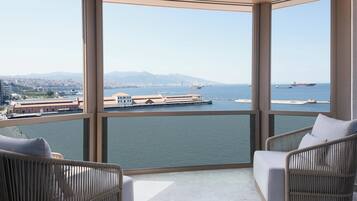 Un balcon avec deux fauteuils en osier, une vue sur un port avec des navires, et un paysage urbain en arrière-plan.