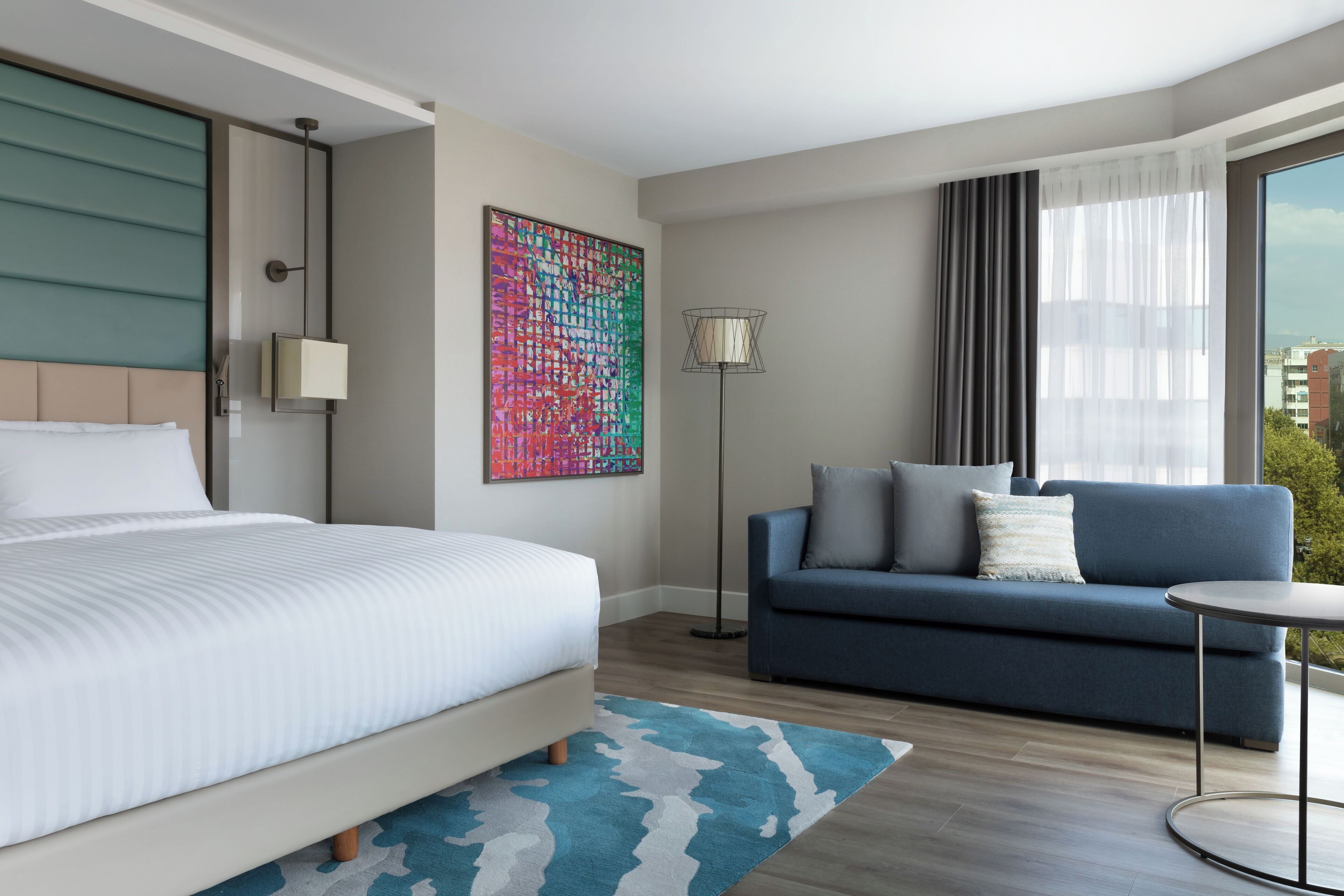 Quarto de hotel moderno com uma cama grande, um sofá, um abajur e uma janela ampla com cortinas.