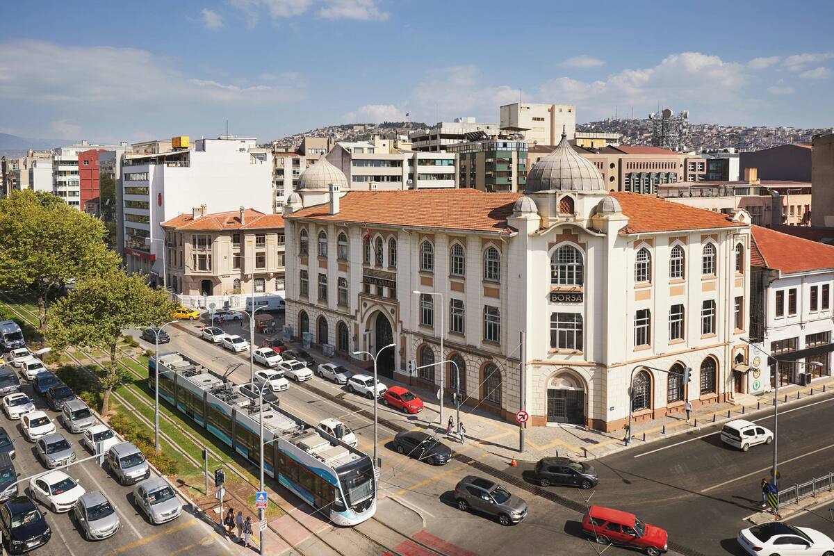 A cityscape with a prominent building featuring arched windows and a dome, surrounded by modern buildings and a tram line.