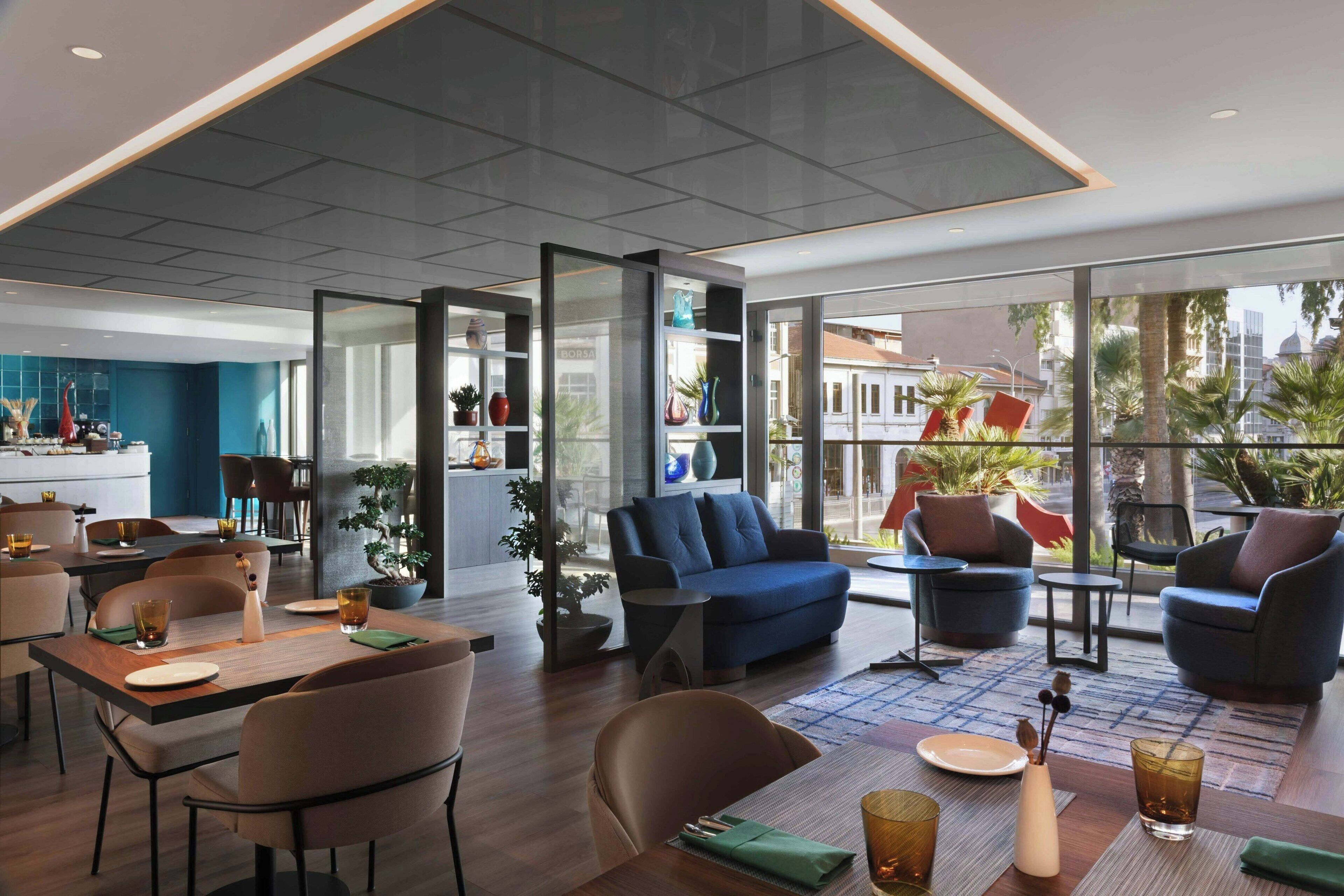 A modern hotel lobby with a dining area, seating, and large windows offering city views.