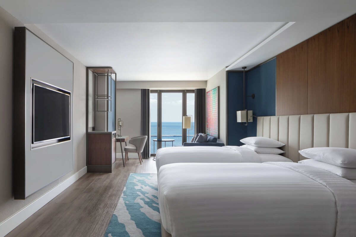 A modern hotel room with two large beds, a desk, a chair, and a view of the ocean.