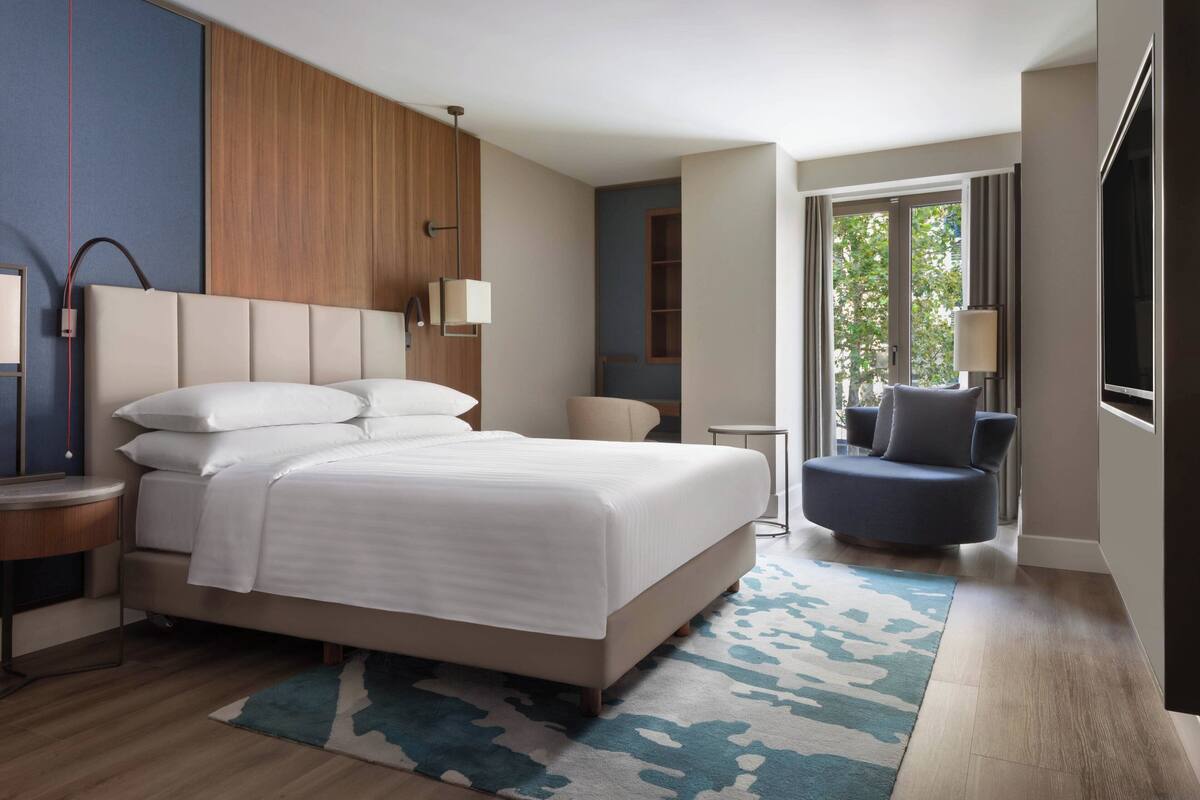 A modern hotel room with a large bed, a seating area, and a view of greenery outside.
