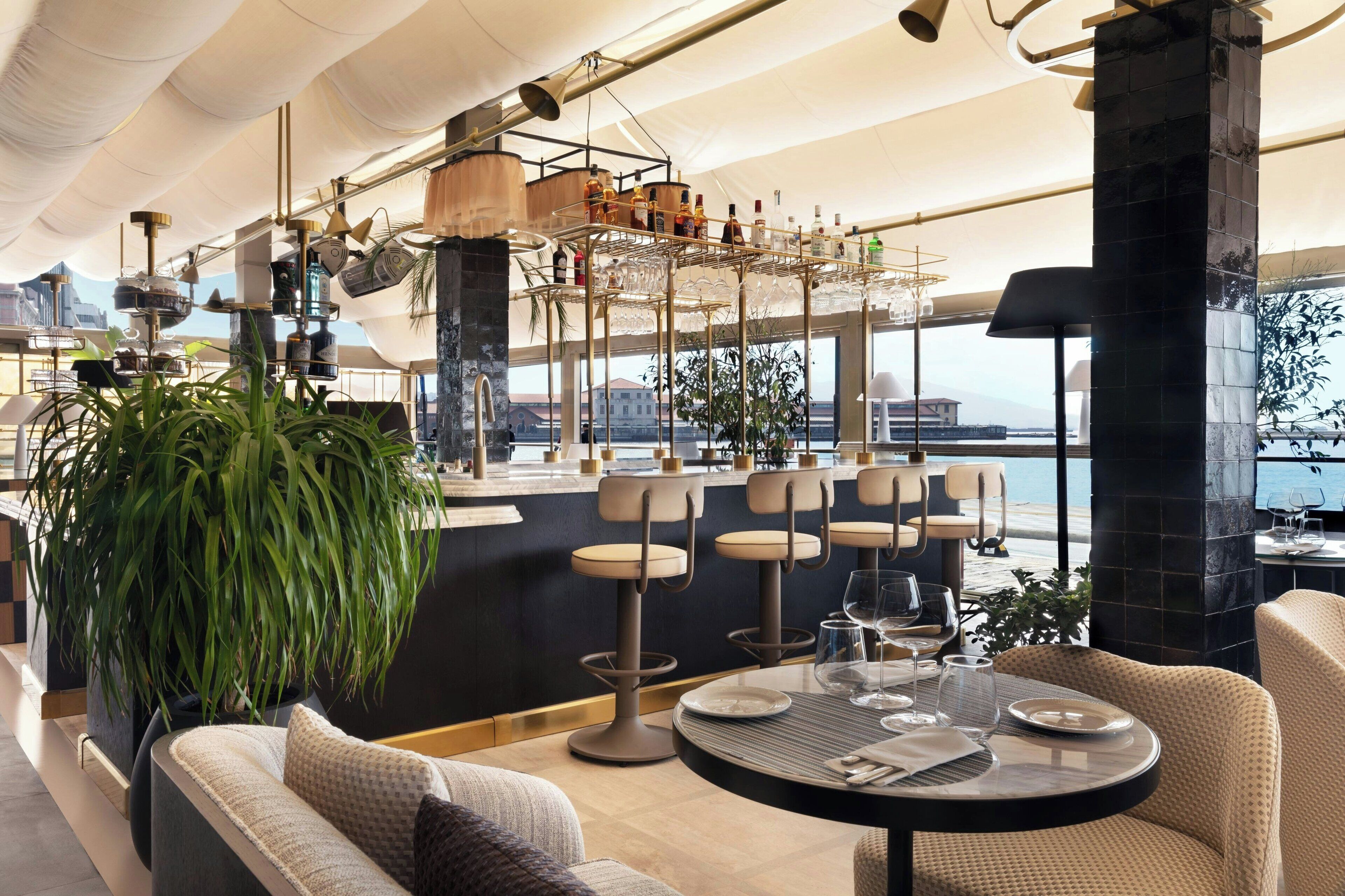 Eine moderne Bar mit mehreren Hockstühlen, einer großen Topfpflanze und Blick auf das Meer.