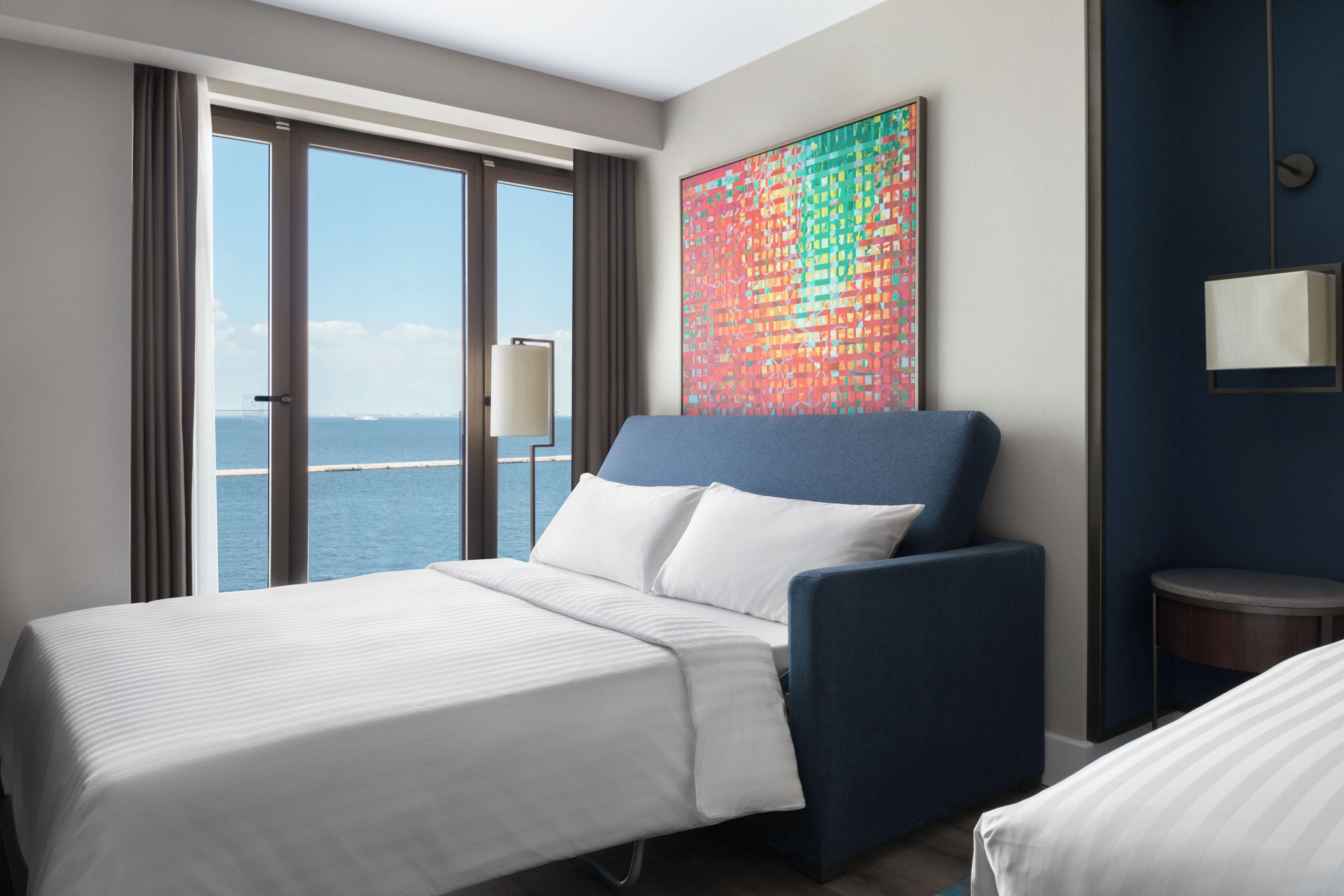 A hotel room with a bed, a blue chair, a view of the sea, and a colorful abstract painting on the wall.