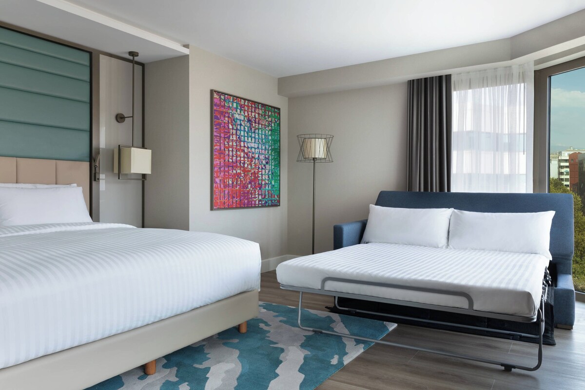 Een moderne hotelkamer met twee bedden, een groot raam met gordijnen, een kleurrijk abstract schilderij en een vloerlamp.