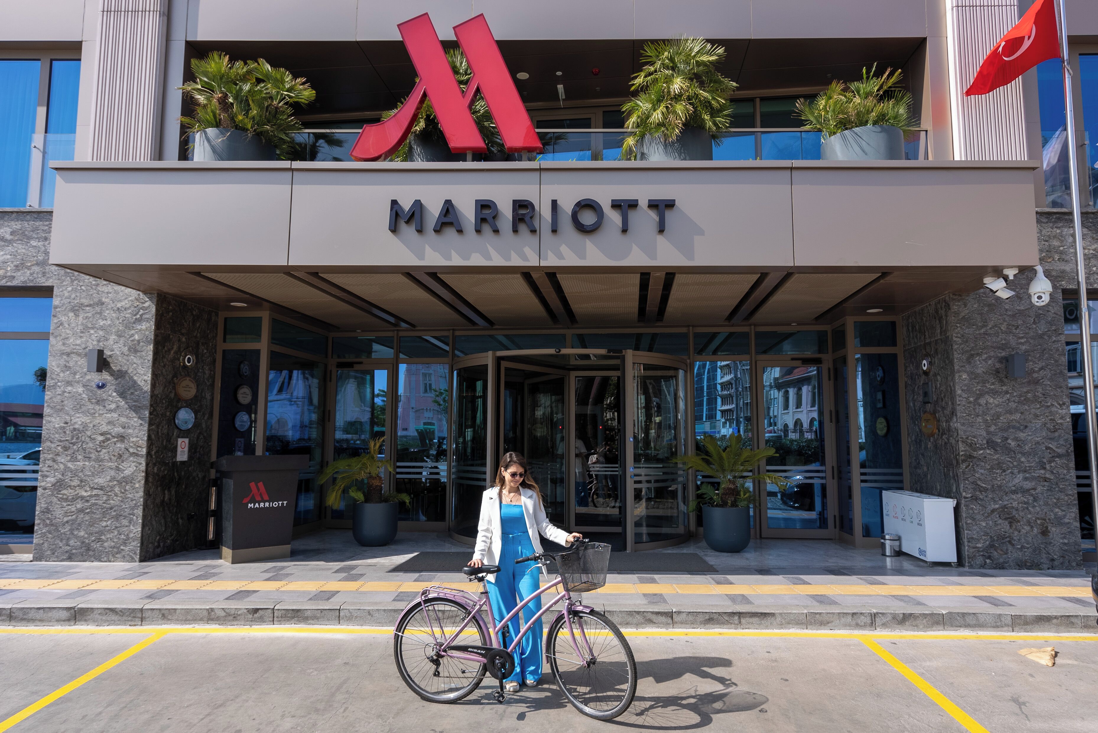 Entrada de un hotel Marriott con un gran logotipo 'M' rojo, plantas en macetas y una persona de pie junto a una bicicleta.