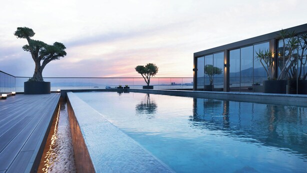 A modern infinity pool with a wooden deck, potted trees, and a glass building with a view of the city skyline at sunset.