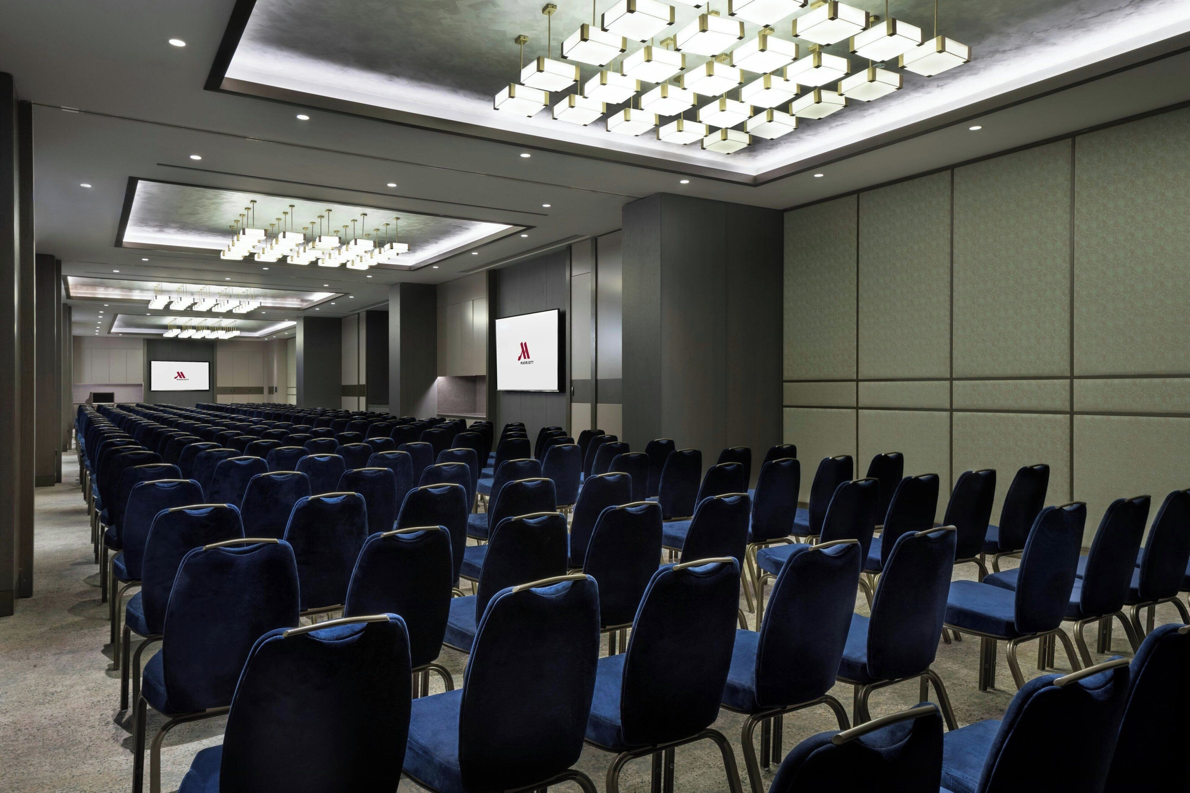 Una sala conferenze con file di sedie blu, un grande schermo e moderne lampade.