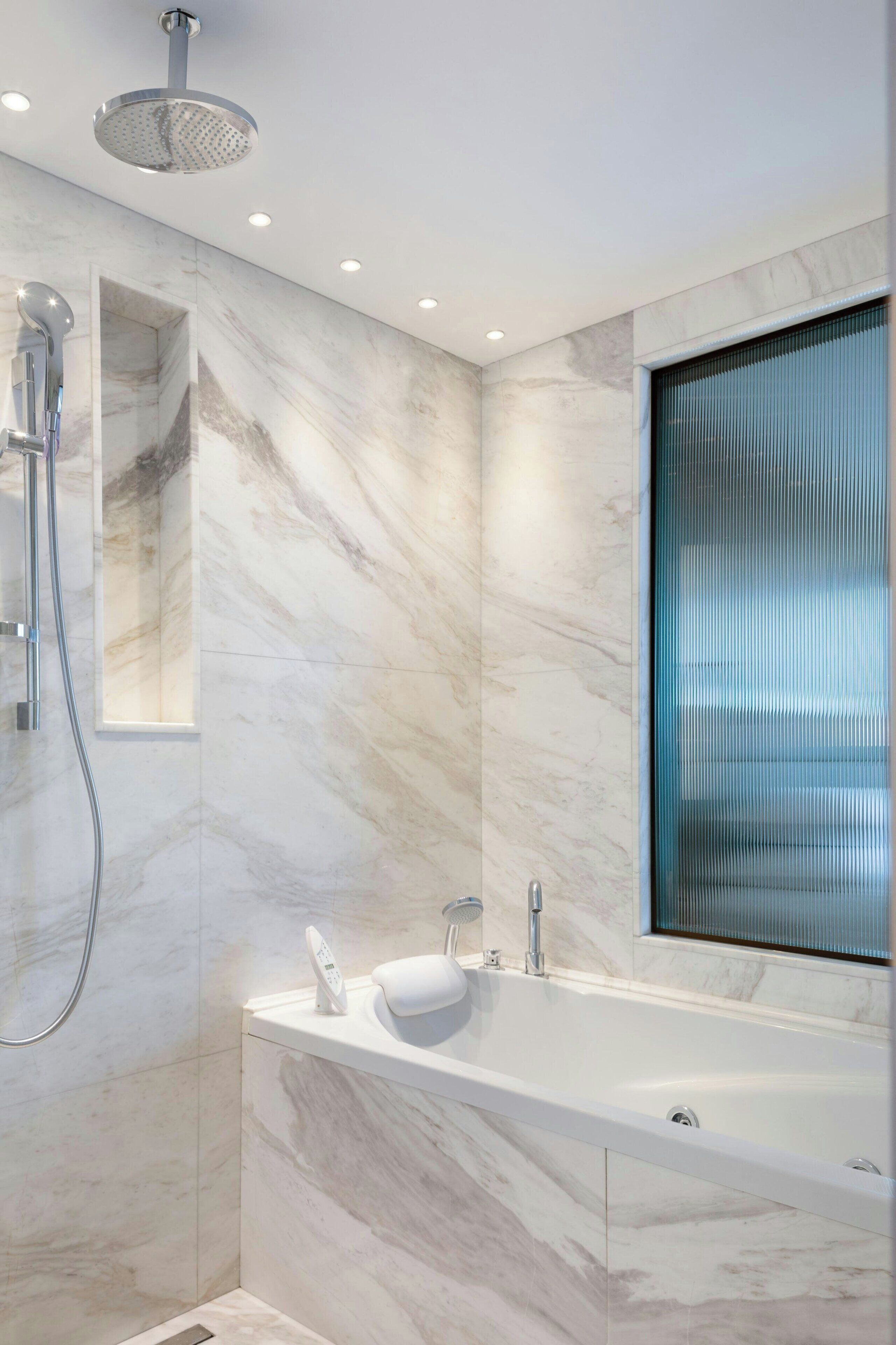Un baño moderno con pared de mármol, una bañera independiente, un cabezal de ducha de mano y un amplio ventanal con persianas.