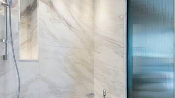 Un baño moderno con pared de mármol, una bañera independiente, un cabezal de ducha de mano y un amplio ventanal con persianas.