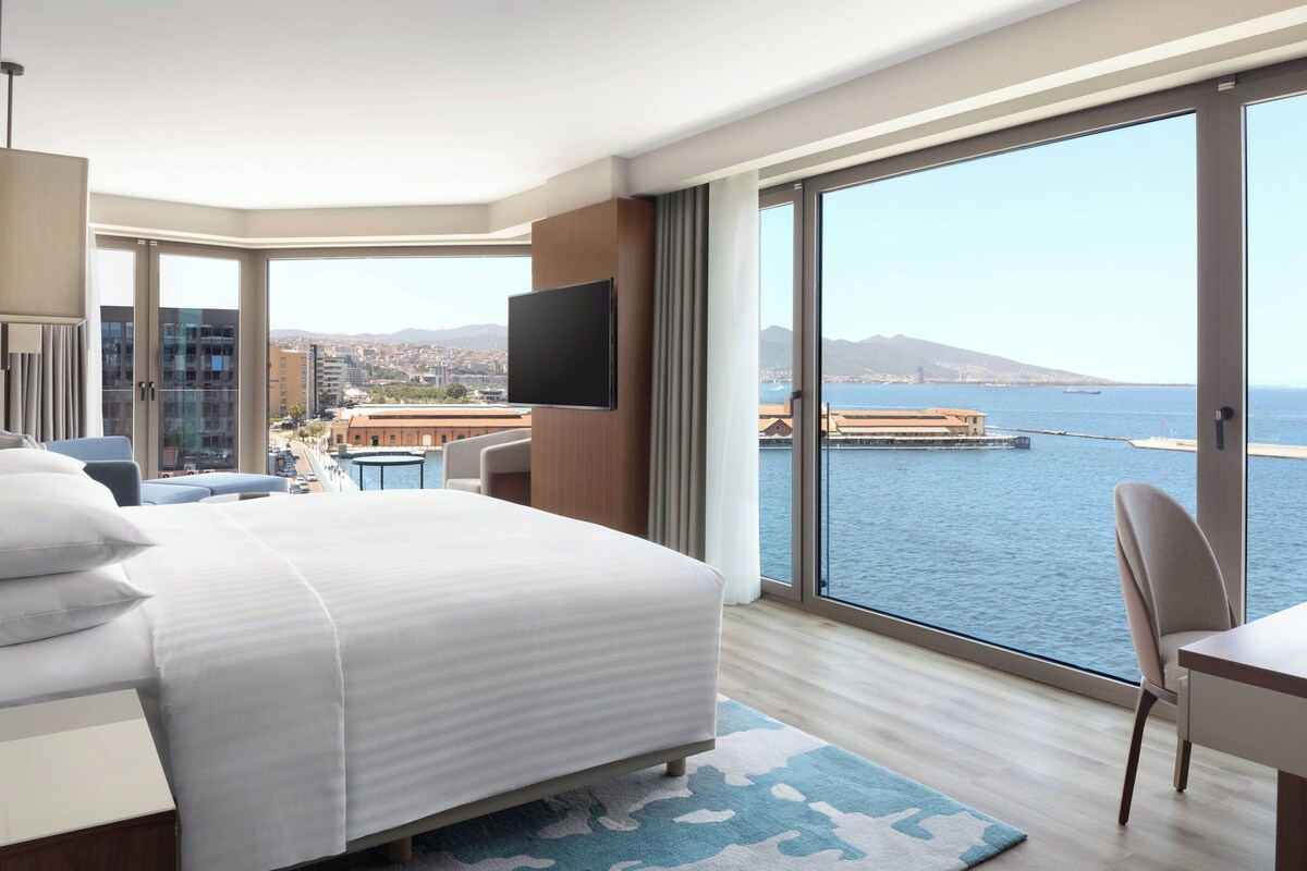 A modern hotel room with a large bed, a desk, and a chair. It features a large window with a view of the sea and a cityscape.