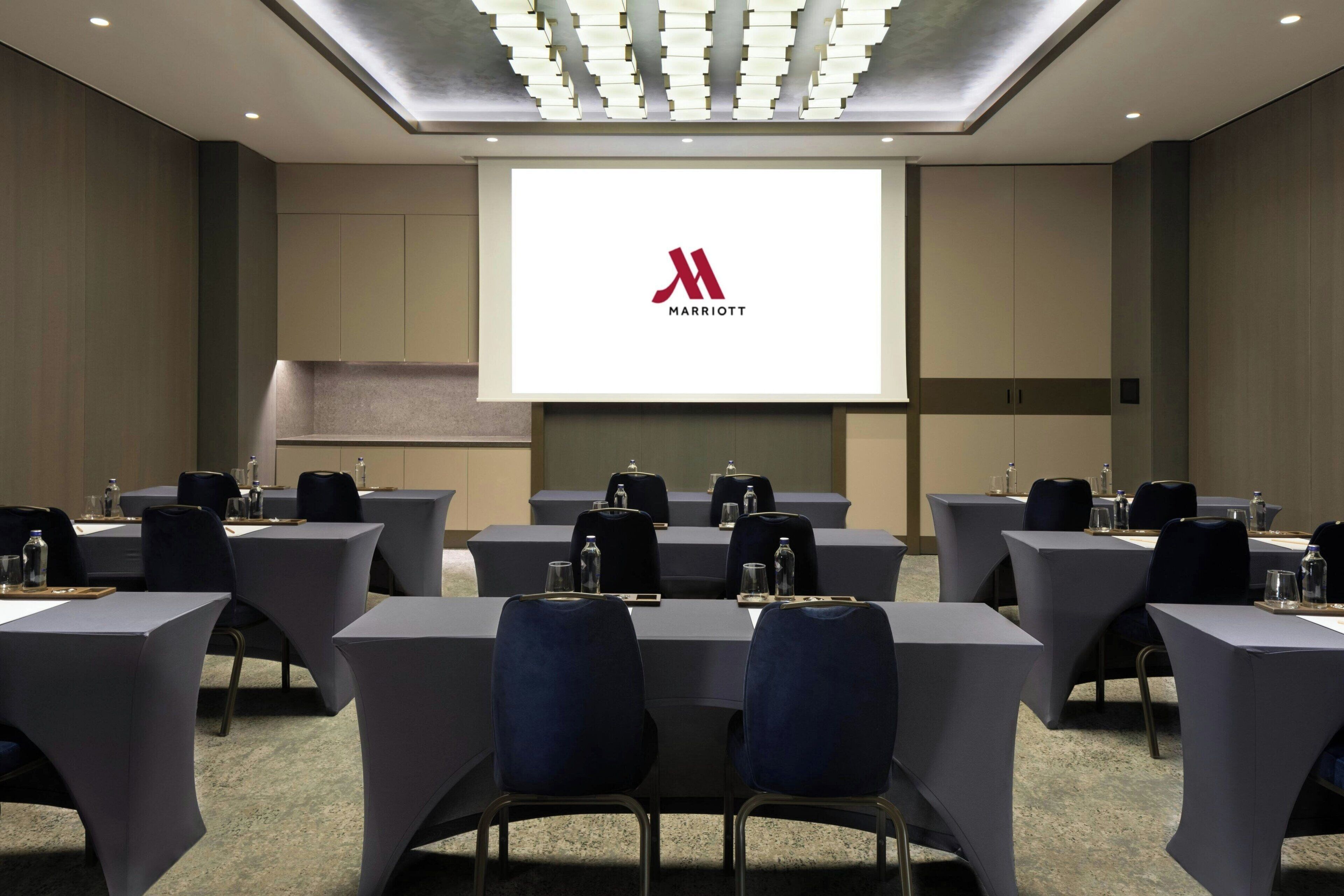 A conference room with a large screen displaying the Marriott logo, arranged tables with chairs, and a modern ceiling design.