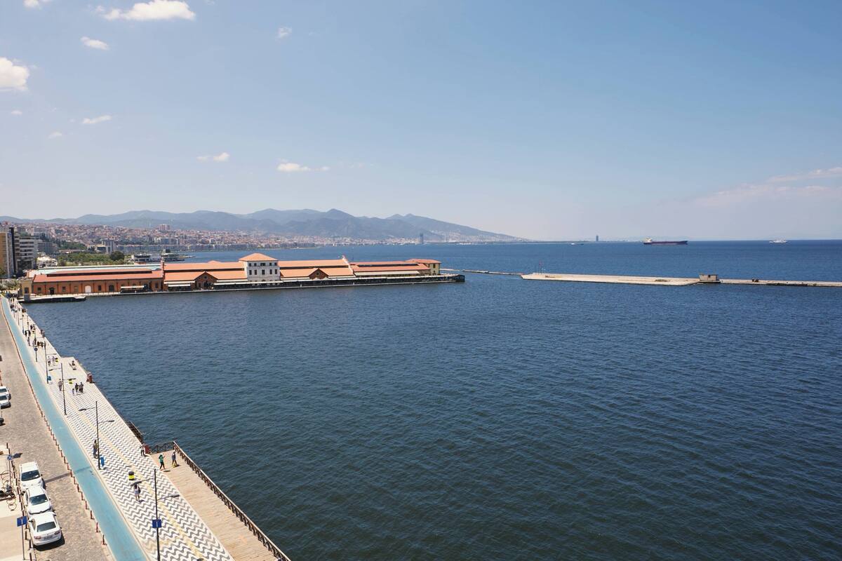 A waterfront promenade with a row of buildings, a pier extending into the water, and a distant mountain range.