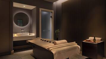 A modern spa room with a massage table, a sink, and a small table with a bottle and bowl.